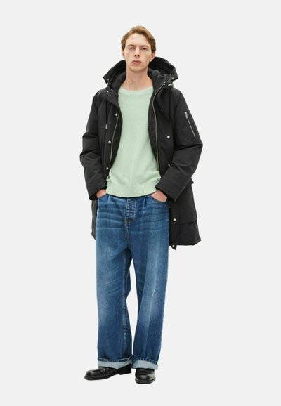 Parka nera con cappuccio sopra un maglione verde chiaro, abbinata a jeans blu larghi e scarpe nere. Tra le caratteristiche ci sono zippers e un tessuto testurizzato.