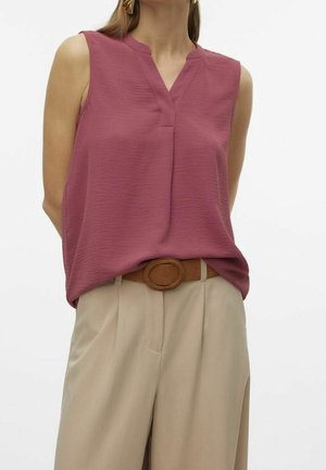 Kobieta nosząca bezrękawnik o fakturze w kolorze mauve, wpuszczony w beżowe spodnie z wysokim stanem z brązowym paskiem z owalną klamrą, na jednolitym tle.