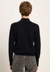 Person mit kurzem braunem Haar, der ein schwarzes Langarmoberteil und hoch taillierte Hosen mit Leopardenmuster trägt, mit dem Rücken zu einer einfarbigen Hintergrund.