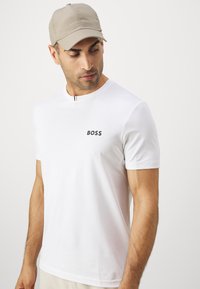 Valge lühikeste varrukatega t-särk musta "BOSS" logoga, ümmargune kaelus ja sile tekstuur. Riietuse täiendab heleda beeži värvi müts.