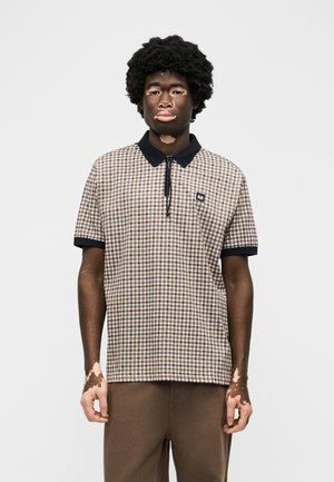 Νεαρός άνδρας με λεύκη φοράει κοντομάνικο πόλο πουκάμισο με καρό σχέδιο και καφέ παντελόνι, στέκεται μπροστά σε απλό λευκό φόντο.