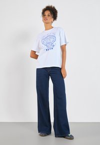 Vit t-shirt med blå snäcka och "BEACH"-grafik, ihop med vida mörkblå jeans. Avslappnad passform, mjuk textur och korta ärmar.