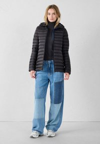 Schwarze Steppjacke mit Kapuze, silbernem Reißverschluss und blauen Akzenten, kombiniert mit hellblauen Patchwork-Jeans und grauen Turnschuhen.