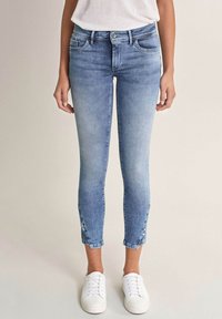 Světle modré džínové kalhoty s těsným střihem, středním pasem a zkrácenou délkou, s knoflíkovými detaily na lemu, spárované s bílými teniskami.