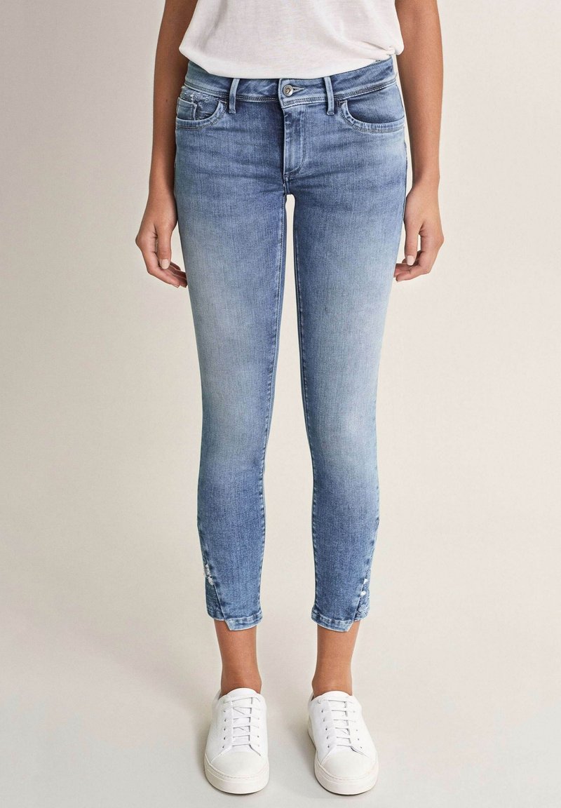 Světle modré džínové kalhoty s těsným střihem, středním pasem a zkrácenou délkou, s knoflíkovými detaily na lemu, spárované s bílými teniskami.