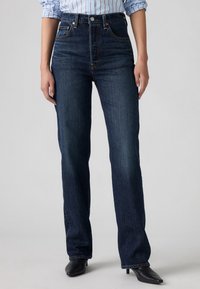 Kvinde iført mørkeblå, højtaljede, lige jeans, stribet skjorte indstukket og sorte ankelstøvler med hæl mod grå baggrund.