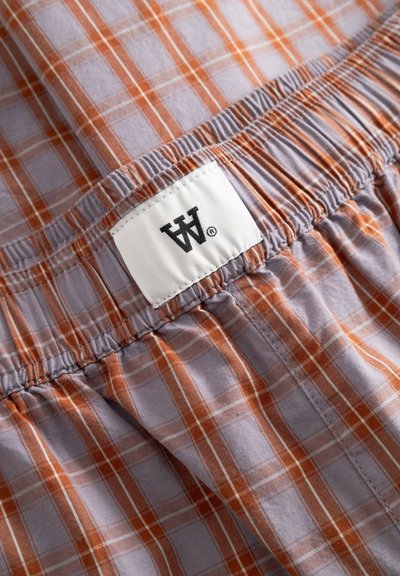 Gros plan sur un tissu à carreaux orange et gris avec une taille élastique et une étiquette blanche portant un logo "W" noir.