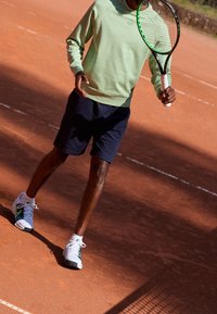 Felpa verde, pantaloni shorts blu navy e scarpe da tennis bianche con dettagli verdi. Tiene una racchetta da tennis su un campo in terra rossa.