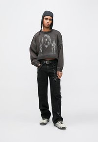 Šedá krátká mikina s kovovým grafickým vzorem, doplněná černými džínovými kalhotami s cvočky. Šedá čepice doplňuje outfit.