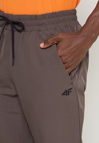 Brązowe spodnie sportowe wykonane z lekkiego materiału, z elastycznym pasem i sznurkiem do regulacji. Posiadają boczne kieszenie i czarny wzór logo.