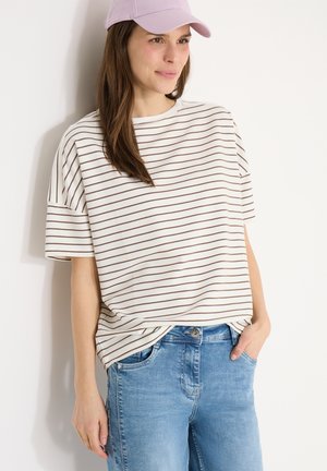 Kobieta w jasnofioletowej czapce, białej koszulce z krótkim rękawem w brązowe paski oraz niebieskich dżinsach, stojąca na tle białej ściany.