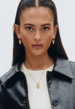 Goldene, herzförmige Ohrringe und passender Anhänger, getragen mit einer schwarzen Lacklederjacke und einem cremefarbenen Pullover, vor einem neutralen Hintergrund präsentiert.