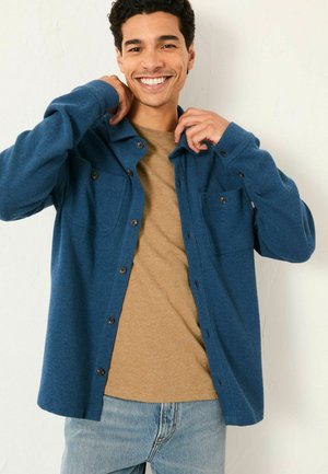 Blaues strukturiertes Button-up-Hemd mit zwei Brusttaschen, getragen über einem sandfarbenen Rundhalsausschnitt-Shirt. Kombiniert mit hellblauen Jeans.