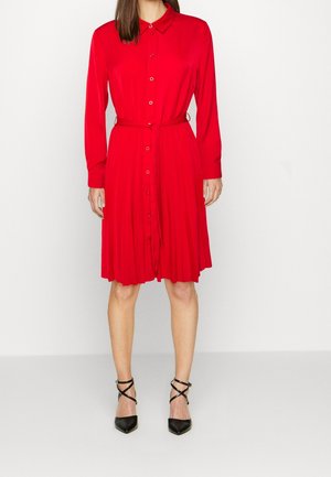 Kobieta ubrana w czerwoną sukienkę na guziki do kolan z długimi rękawami i paskiem w talii, w połączeniu z czarnymi szpilkami z paskami krzyżującymi się na palcach.