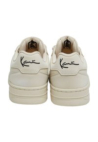 Bežové tenisky s hladkým koženým svrškem, kulatou špičkou, silnými podrážkami a černým podpisovým detailem na patě. Texturovaná podšívka uvnitř.