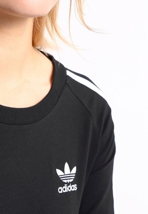 Czarna bluza z okrągłym dekoltem, białe paski na ramionach oraz haftowane białe logo Adidas z przodu. Miękka, teksturowana tkanina.