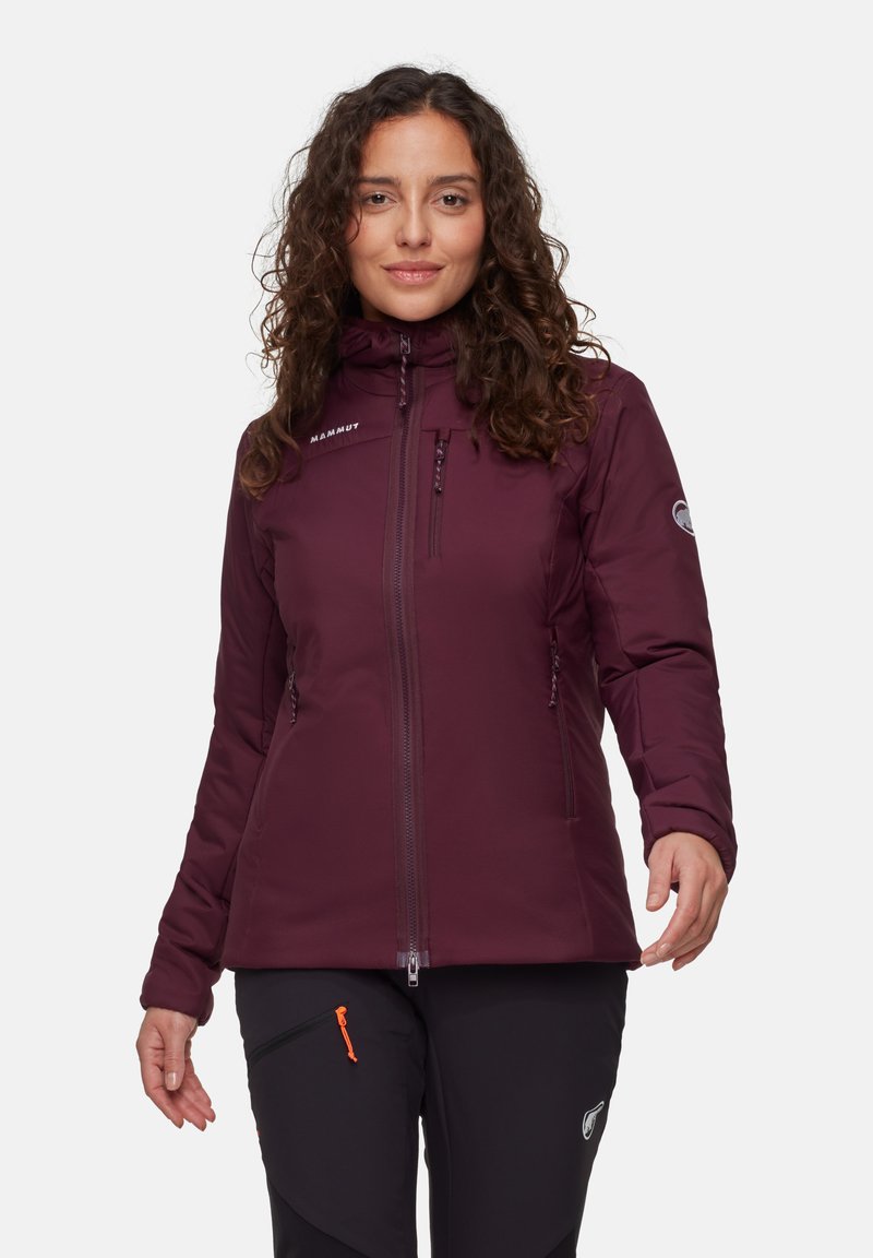 Vrouw met krullend haar, gekleed in een bordeauxrode zip-up jas en zwarte broek met een oranje ritsdetail, staande voor een effen witte achtergrond.
