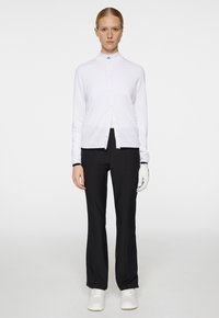 Weißer Knopf-Cardigan über einem schwarzen Rollkragenpullover, kombiniert mit schwarzen Hosen und weißen Sneakern. Accessoire: Ein weißer Handschuh an einer Hand.