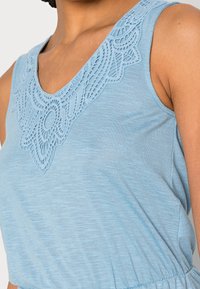 Top sin mangas de color azul claro, con un escote en V y un intrincado detalle de encaje alrededor del cuello. Tejido suave y texturizado, y cintura elástica.