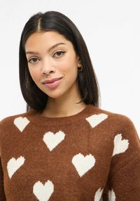 Hnědý pletený svetr s bílým vzorem srdcí, kulatým výstřihem a texturovanou látkou. Měkký materiál vytváří útulný vzhled.