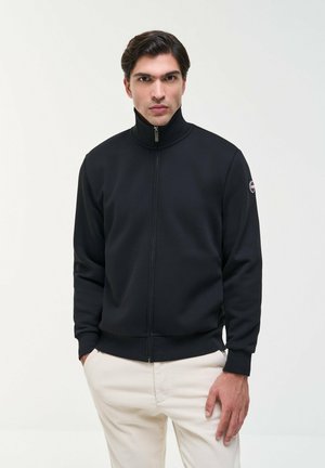 Homme portant une veste noire zippée avec un col montant et un pantalon clair, se tenant devant un fond blanc uni.