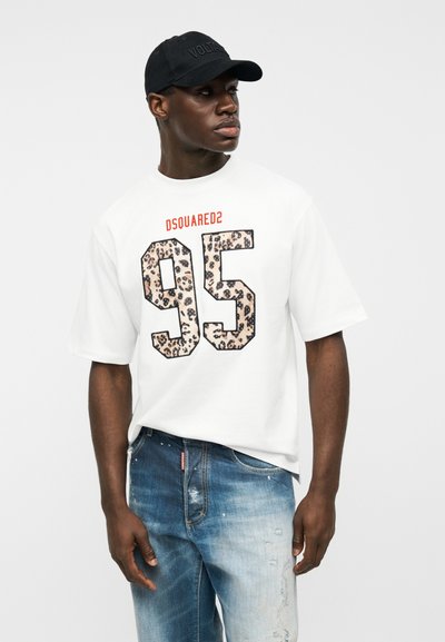 Άνδρας που φοράει λευκό μπλουζάκι με το κείμενο "DSQUARED2 95" σε λεοπάρ σχέδιο και ξεβαμμένο μπλε τζιν, κοιτάζει προς τα δεξιά, φορώντας μαύρο καπέλο.