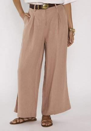 Pantalon marron à jambes larges avec plis, porté avec une ceinture marron, des bijoux dorés, un haut blanc et des sandales plates marron à brides sur fond clair.