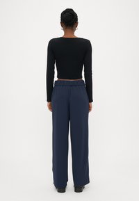 Kvinne med flette, iført sølv hoop-øredobber, svart langermet croptopp, marineblå vide benbukser og svarte sko, stående med ryggen mot.