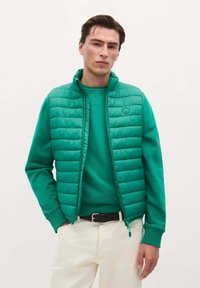Chaleco acolchado verde con cuello alto, cierre de cremallera y patrón texturizado, combinado con una sudadera verde a juego y pantalones crema.