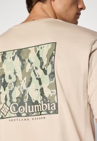 Beige t-skjorte med et stort grønt og beige kamuflasjemønster på baksiden, med "Columbia" logo og teksten "Portland, Oregon" nedenfor.