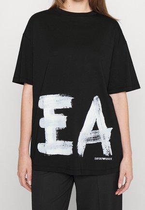Persoon draagt een zwarte oversized T-shirt met grote witte geschilderde letters "EA" en een klein Emporio Armani-logo rechtsonder.