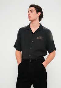 Μαύρο πουκάμισο με κοντά μανίκια από απαλό ύφασμα, με γιακά και διακριτικό λογότυπο. Συνδυάζεται με υφασμένα μαύρα βελούδινα παντελόνια.