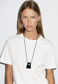 Maglietta bianca in cotone con scollo rotondo, indossata con una collana con ciondolo geometrico nero su cordino nero. Sfondo uniforme.