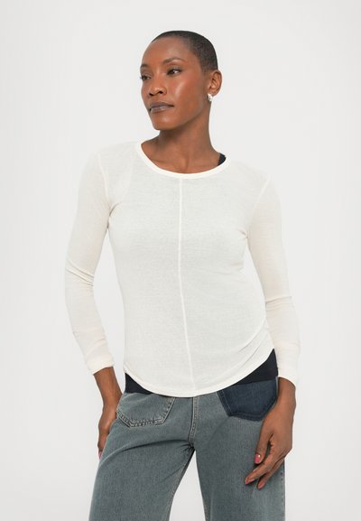 Long-sleeve, light cream shirt with a rounded neckline and textured fabric, paired with high-waisted denim jeans featuring contrasting patches.