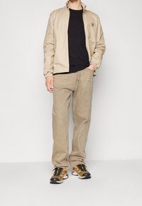 Veste beige à col montant, pull noir, pantalon en velours côtelé beige, et baskets multicolores avec des accents noirs.