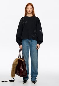 Sudadera negra con detalles joya, combinada con jeans de tiro alto en azul claro. Bolso tote burdeos oscuro con detalle de piel sintética. Zapatos negros puntiagudos.