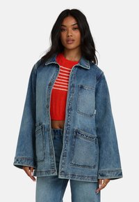 Veste en denim bleu clair, dotée de deux grandes poches avant, avec fermeture par boutons et manches larges. Portée par-dessus un pull rayé rouge.