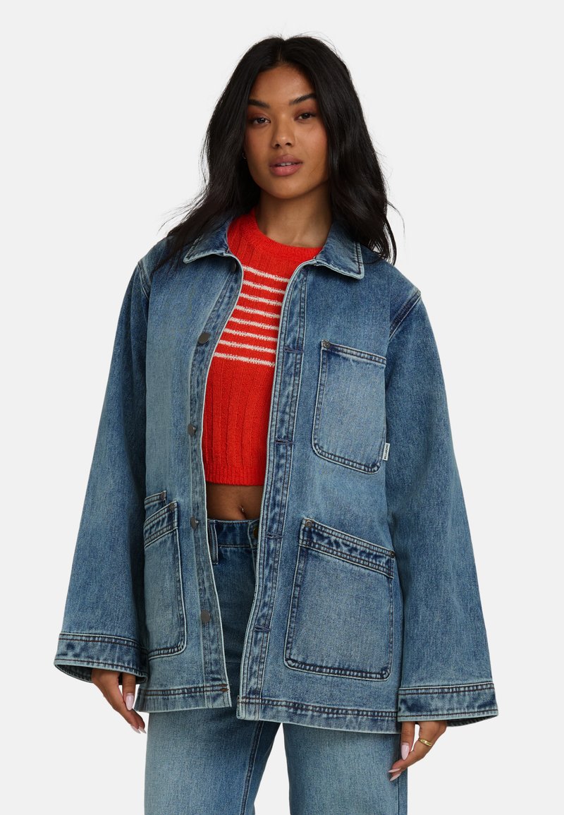 Veste en denim bleu clair, dotée de deux grandes poches avant, avec fermeture par boutons et manches larges. Portée par-dessus un pull rayé rouge.
