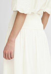 Robe blanche texturée avec une taille smockée et une jupe fluide, présentée de côté, mettant en valeur la légèreté et la douceur du tissu.