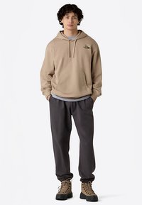 Beige huppartröja med framficka, grått innerlager, kombinerad med mörkgrå avsmalnande byxor och bruna snörstövlar.