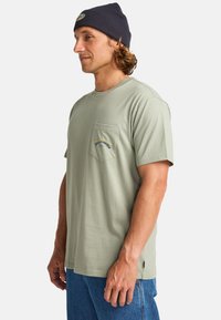 Mintgrøn bomulds-T-shirt med en brystlomme og et blåt broderet logo. Matchet med blå denimjeans og en marineblå beanie.