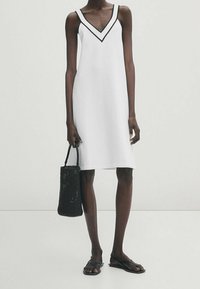 Weißes ärmelloses Kleid mit V-Ausschnitt und schwarzem Besatz. Dazu eine schwarze geflochtene Tasche und flache braune Sandalen. Glatte Stoffstruktur.