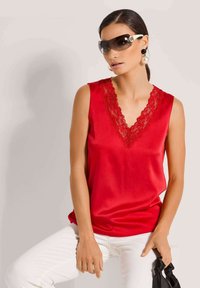 Rotes Satin-Top ohne Ärmel mit einem Spitzen-V-Ausschnitt, kombiniert mit weißen Hosen. Das Modell trägt dunkle Sonnenbrillen und hat ein schwarzes Accessoire.