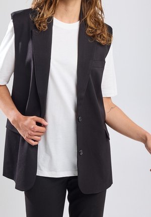 Persoon die een wit T-shirt draagt onder een zwart mouwloos colbert, met één hand die het colbert rechtzet en een kleine golf-tatoeage zichtbaar op de arm.