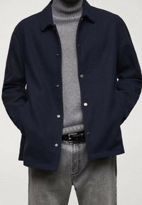 Navy blue wool jacket with snap buttons, classic collar, worn over a gray knitted turtleneck, paired with gray jeans and a black belt.