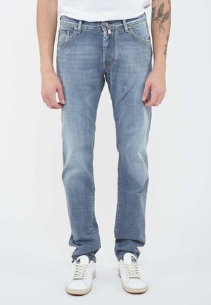 Light blue faded straight-leg jeans worn with white sneakers, showing part of tattooed forearm and hands relaxed at sides.