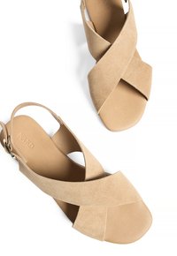 Beige mokkanahka slingback-sandaali, jossa on ristikkäiset hihnat, avoin varvas ja tasapohja. Sen sisäpinnassa on hienovarainen logo ja säädettävä nilkkasolki.
