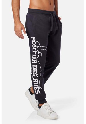 Pantaloni della tuta neri con stampa grafica bianca sulla gamba sinistra, dotati di una vita elastica e caviglie a coste. Indossati con scarpe da tennis bianche.