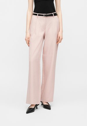 Donna che indossa pantaloni rosa chiaro a gamba larga, cintura nera, top nero senza maniche e tacchi neri a punta su sfondo bianco.