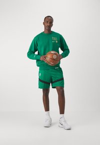 Žalias megztinis su "Boston Celtics" ir Nike logotipu, derinamas su žaliais šortais su juodais akcentais, laikantis krepšinio kamuolį, baltais batais.